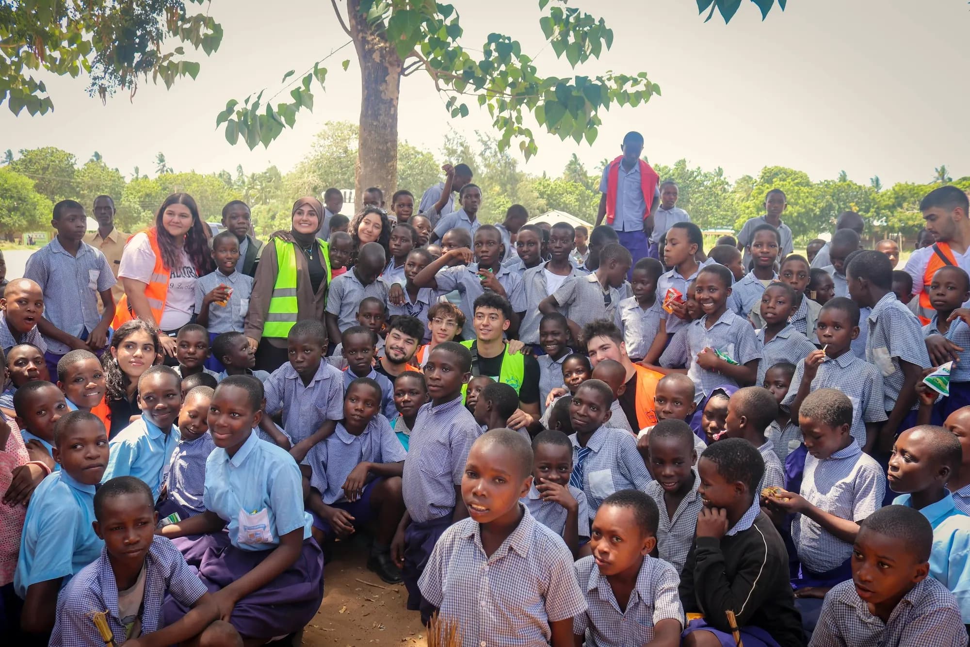 About a hundred Kenyan schoolchildren in blue uniforms sit with LeadUp volunteers under a tree.