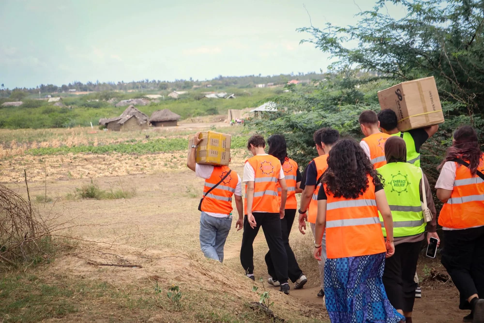 LeadUp volunteers in orange vests carry boxes of solar kits along a dirt path toward a village.